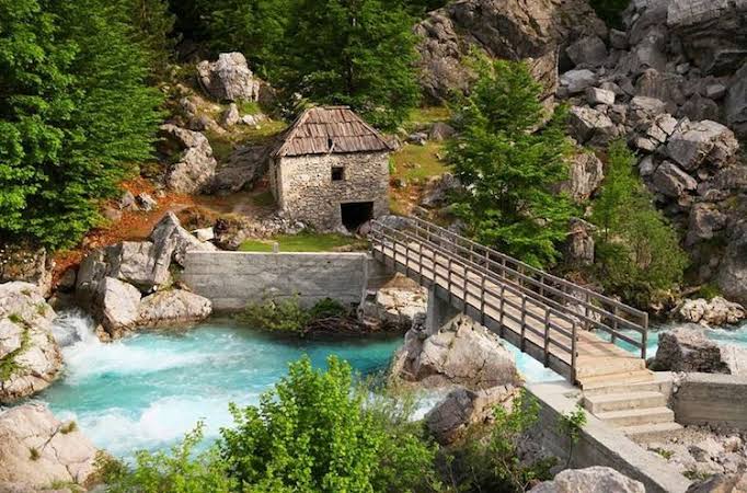 A stone building and wooden bridge beside a bright blue river